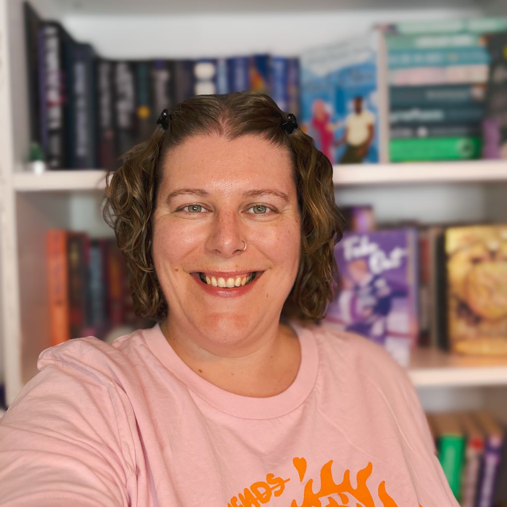 A head shot of author Lowri Charles, a white woman with brown, wavy, shoulder length hair. She is smiling and standing in front of a bookshelf.